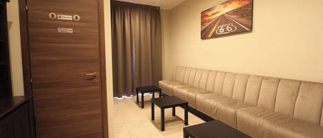 A hotel room with a sofa, two small tables, a door with a sign indicating a toilet and a room number, and a painting of a road on the wall.