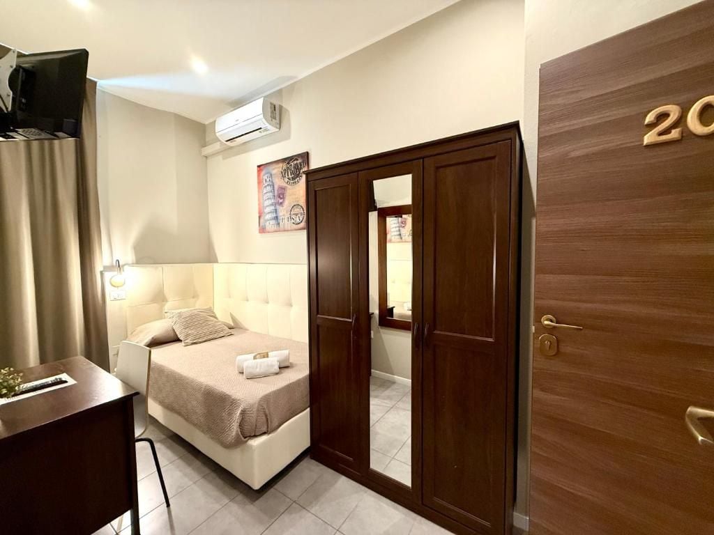 A hotel room with a bed, a desk, an air conditioning unit, a wardrobe, and a door with a number.
