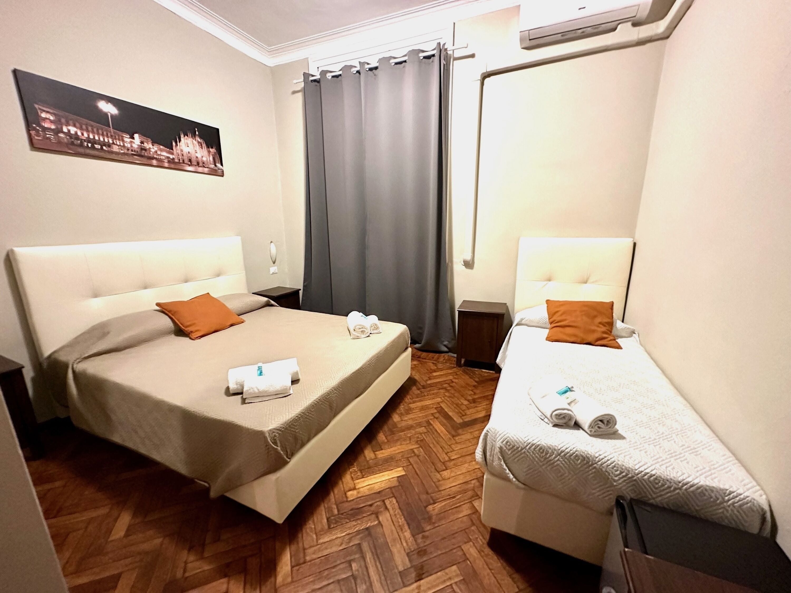 A hotel room with two beds, a wooden herringbone floor, a cityscape painting, and a window with curtains.