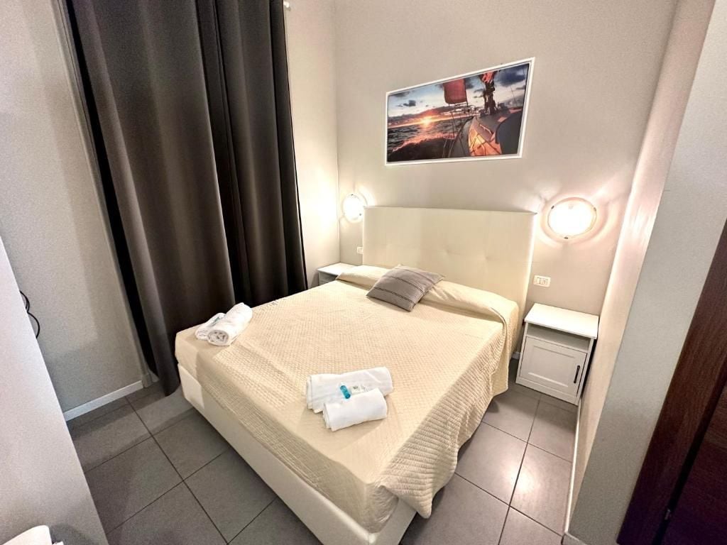 Una cama bien hecha con cubrecolcha color beige, una almohada y dos toallas enrolladas. Se aprecia una mesita de noche con una lámpara y un cuadro colgado en la pared.