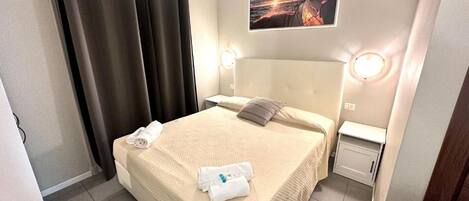 Una cama bien hecha con cubrecolcha color beige, una almohada y dos toallas enrolladas. Se aprecia una mesita de noche con una lámpara y un cuadro colgado en la pared.