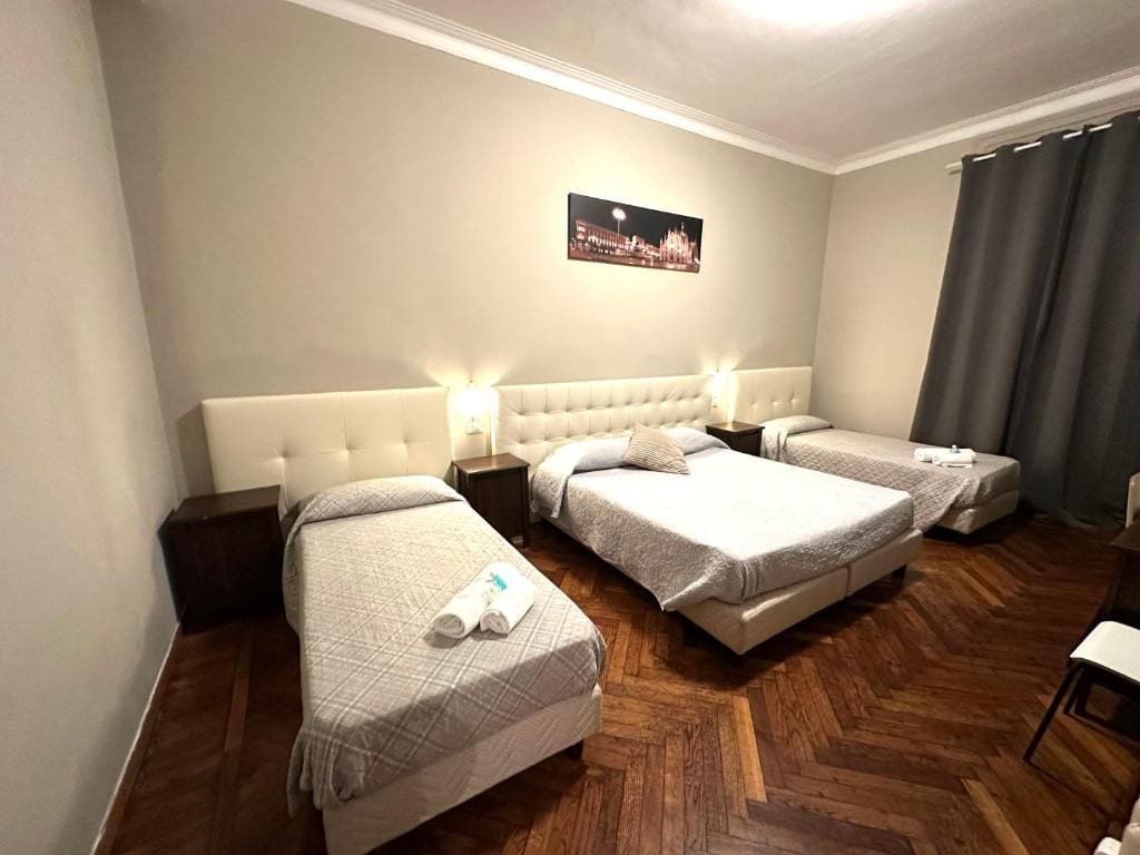 Una camera d'albergo con tre letti, un'ampia finestra con tende e un quadro con vista sulla città.
