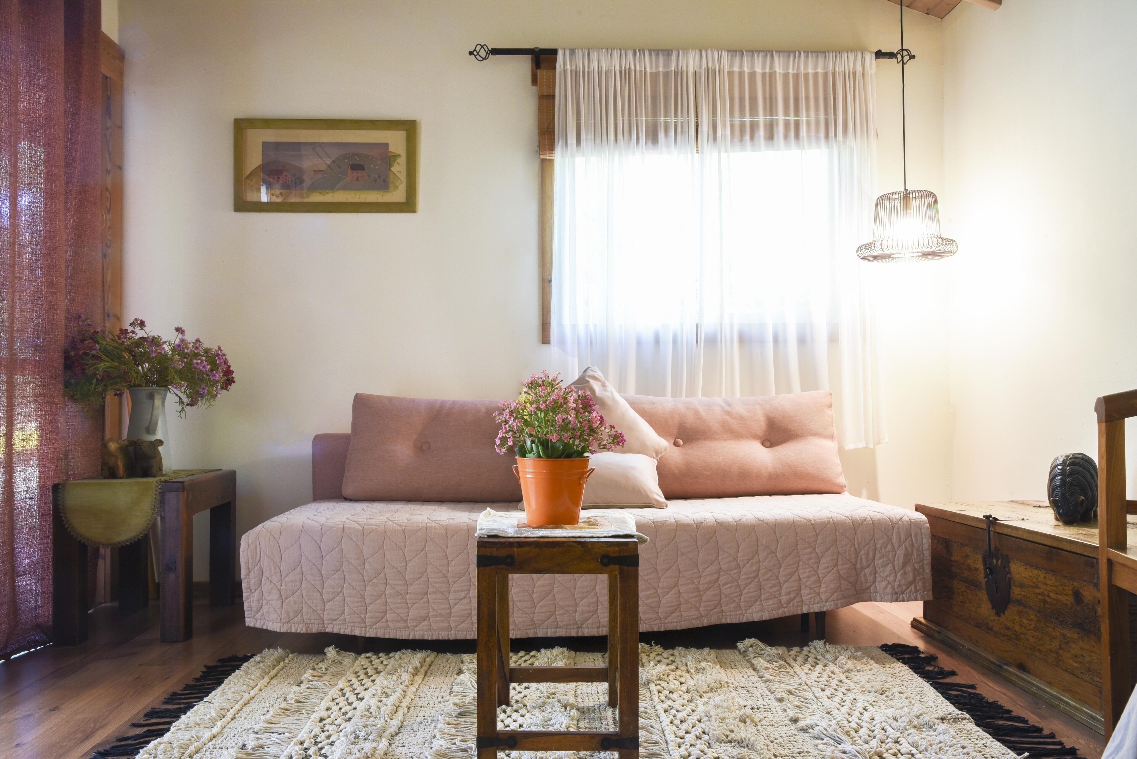 A cozy bedroom with a bed, a small table with a potted plant, a wooden dresser, and a window with sheer curtains.
