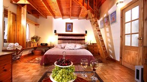 Quarto aconchegante com teto de madeira, cama com colcha florida, uma cesta de frutas e dois copos de vinho.