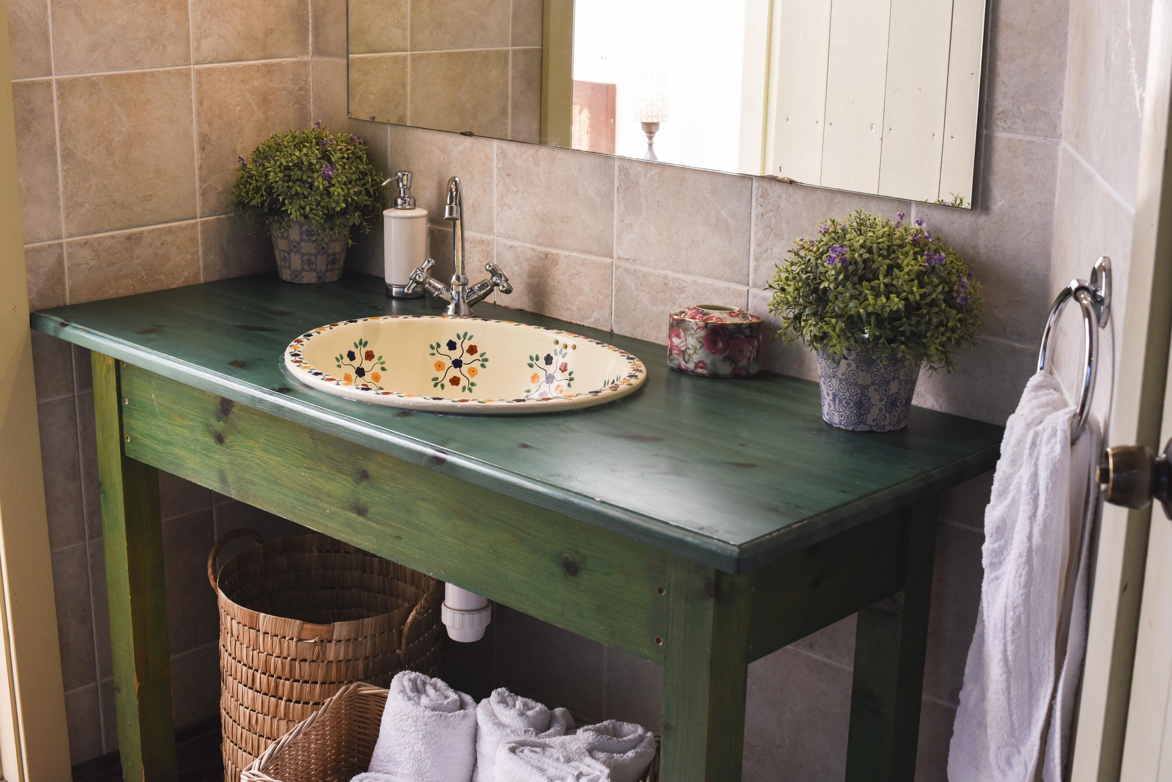 A bathroom with a green vanity, a sink with floral designs, two potted plants, a white towel, and a mirror above the sink.