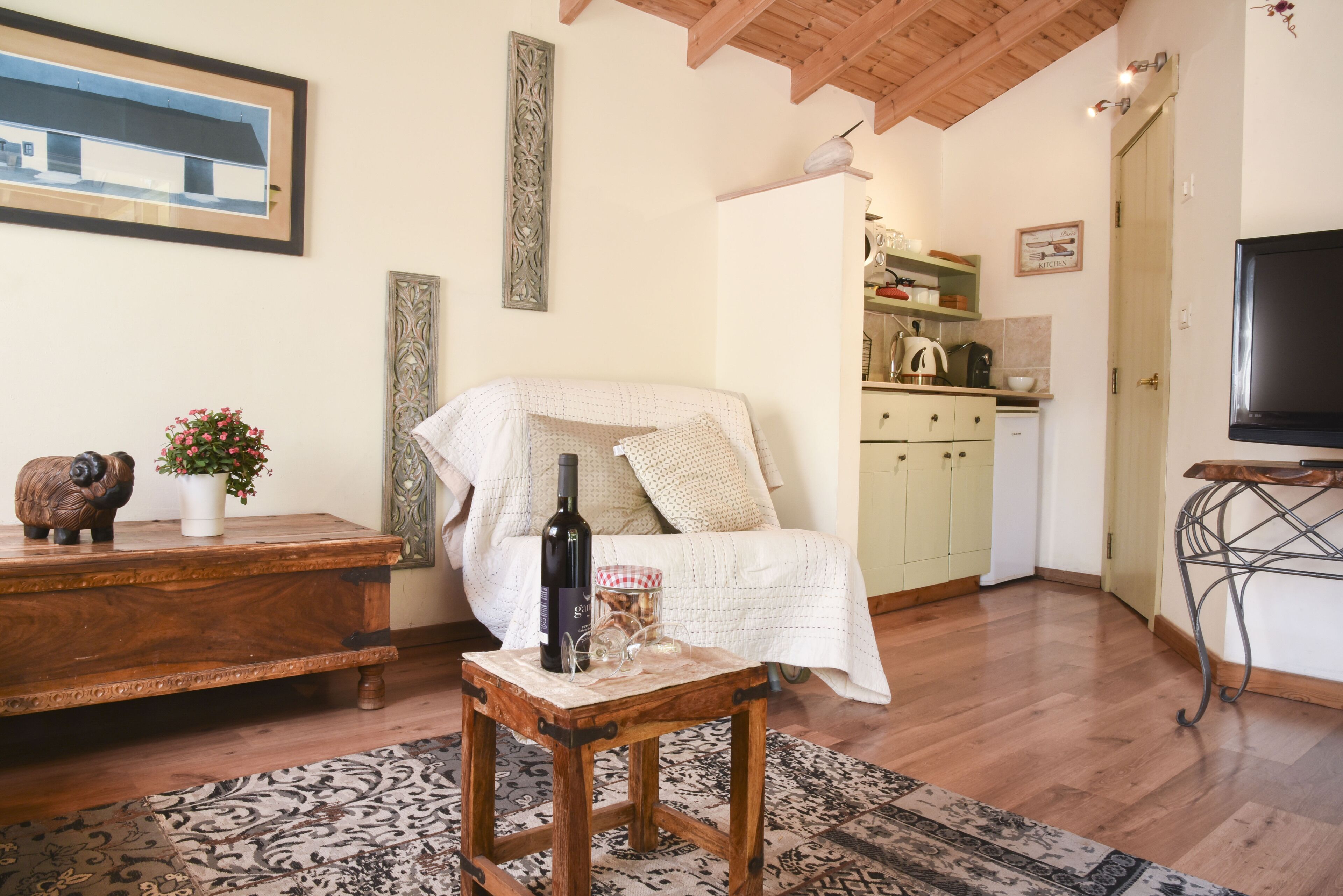 Una habitación acogedora con una cama, una mesa de madera con una botella y un vaso, un tapete y una zona de cocina al fondo.