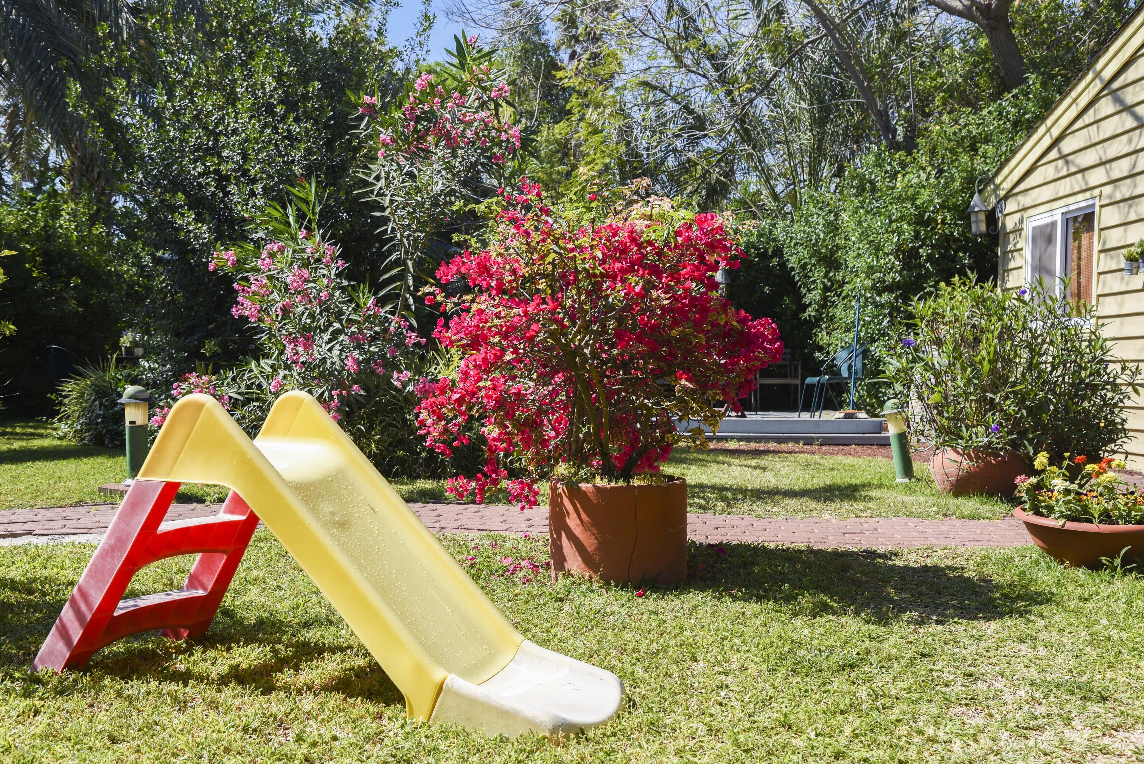A playground with a yellow slide and a red climbing structure, surrounded by lush greenery and flowering plants.