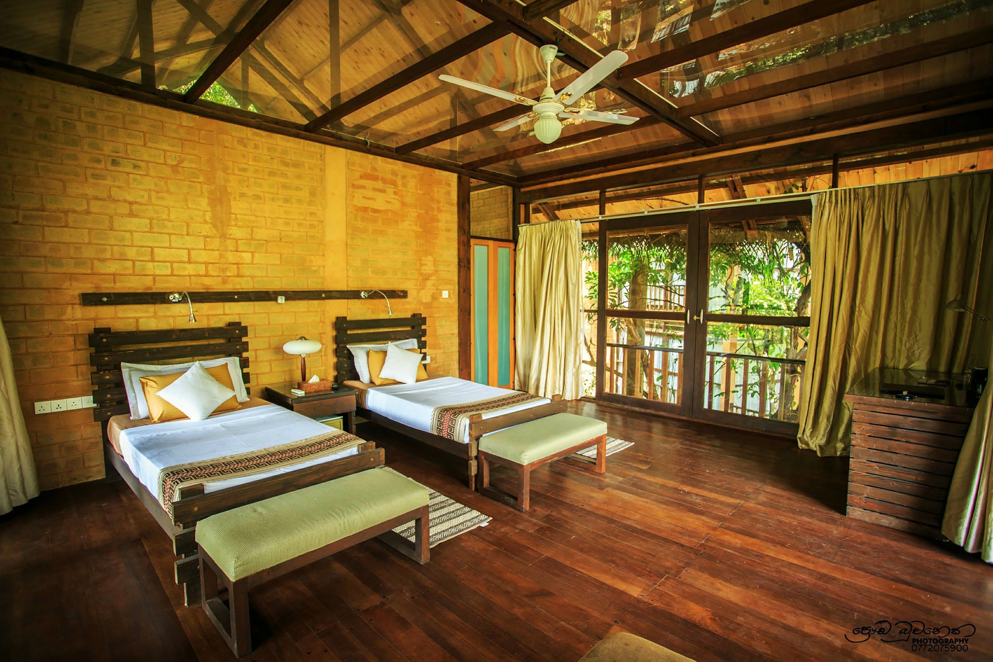 A room with two beds, wooden flooring, a ceiling fan, and a large window with curtains.