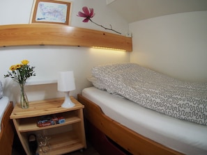 Una habitación pequeña con una cama individual, una mesita de noche con lámpara, un estante con libros y vasos, y un reloj en la pared.
