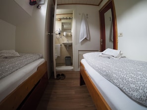 Una habitación pequeña y limpia con dos camas, piso de madera, baño con lavamanos y espejo, y una puerta que da a otra habitación.