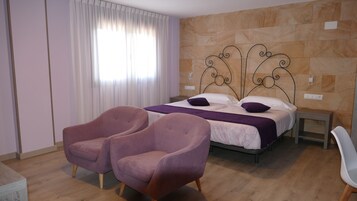 Un dormitorio con una cama, dos sillones, una mesita de noche y un ventanal con cortinas.