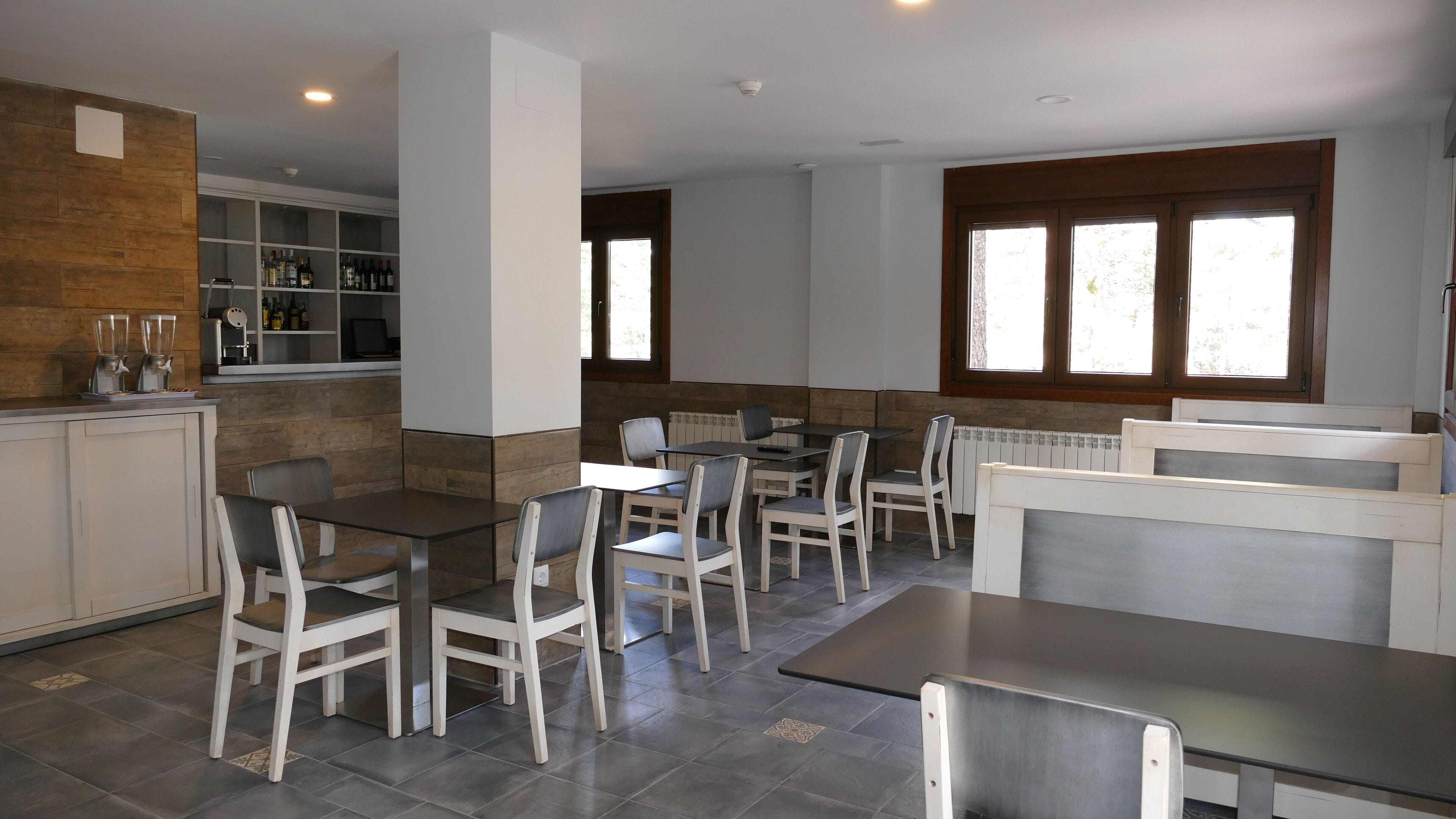 Un comedor moderno con sillas y mesas blancas, una zona de cocina con botellas de vino, y una ventana con luz natural.