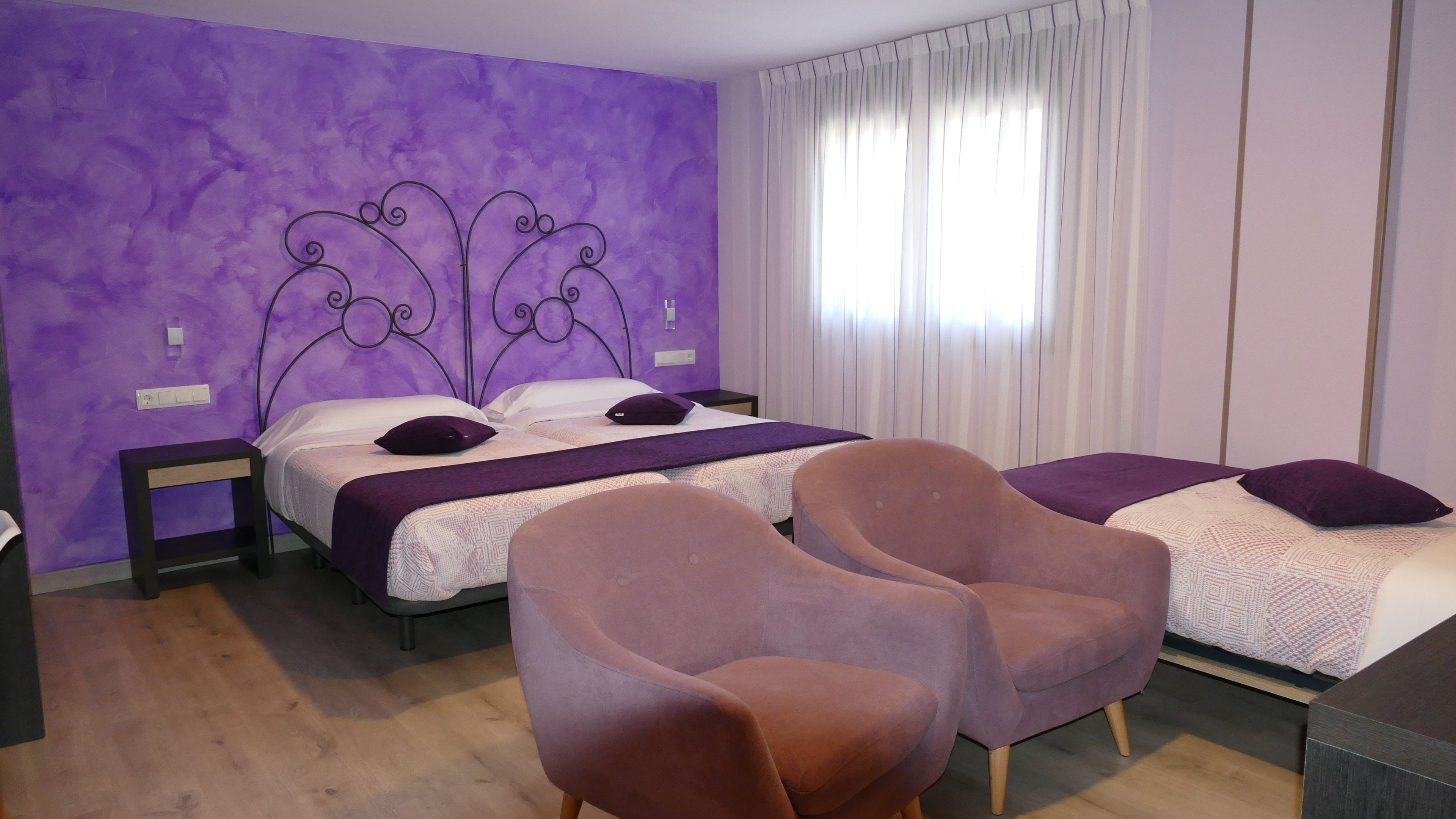 A bedroom with two beds, a purple wall, and a large window with curtains.