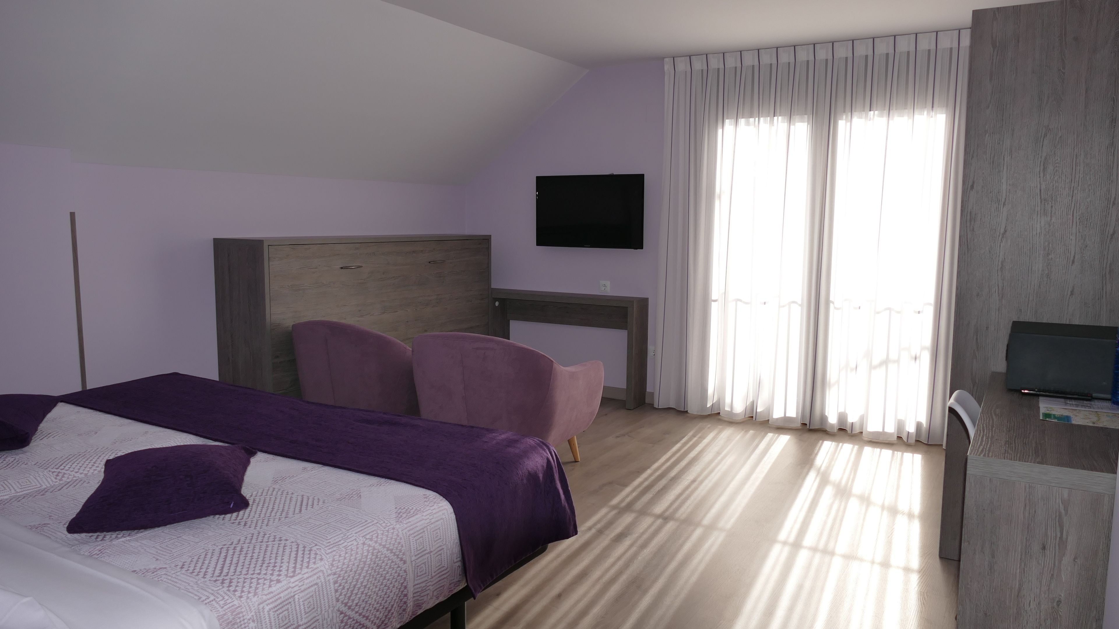 Habitación de hotel con cama, escritorio, silla, televisor y ventana con cortinas.
