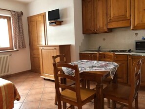 A kitchen with wooden cabinets, a dining table with chairs, a television mounted on the wall, and a window with curtains. - Oberteil (Gressoney-la-Trinite)