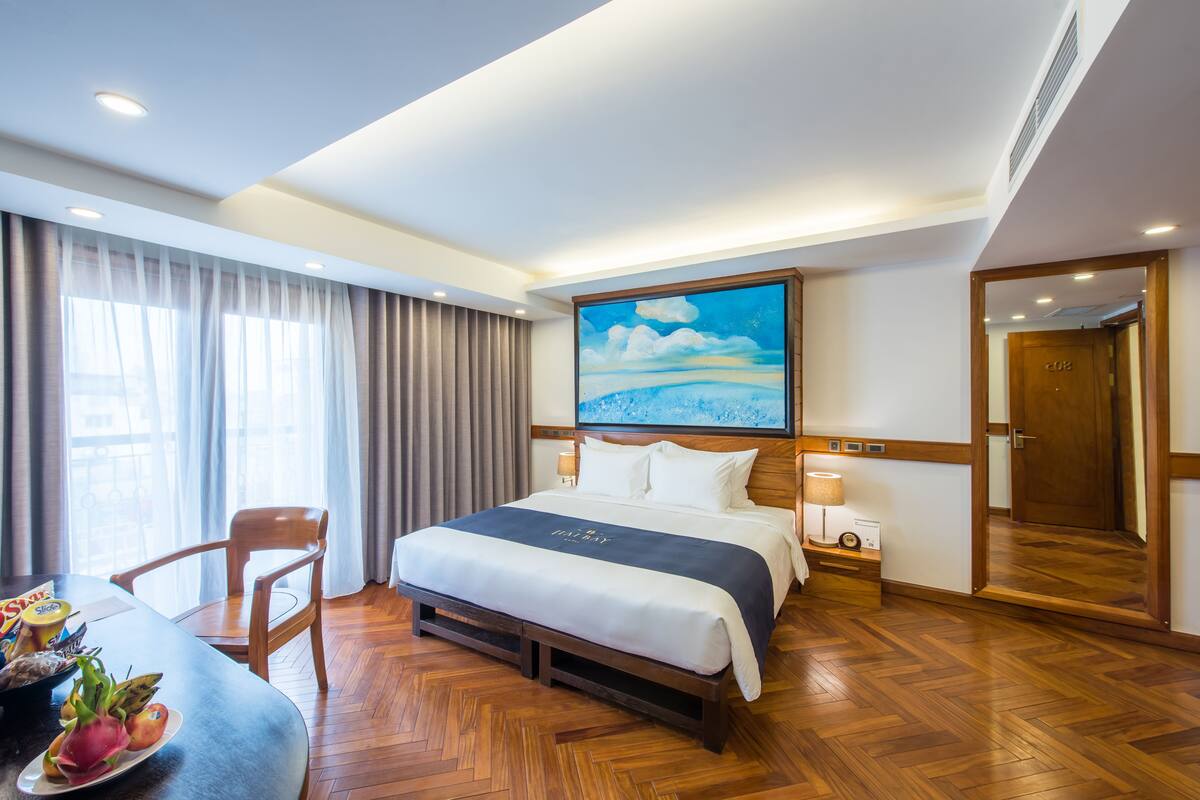 A hotel room with a large bed, a wooden headboard, a painting of a landscape, a desk with a chair, and a dining table with fruit.