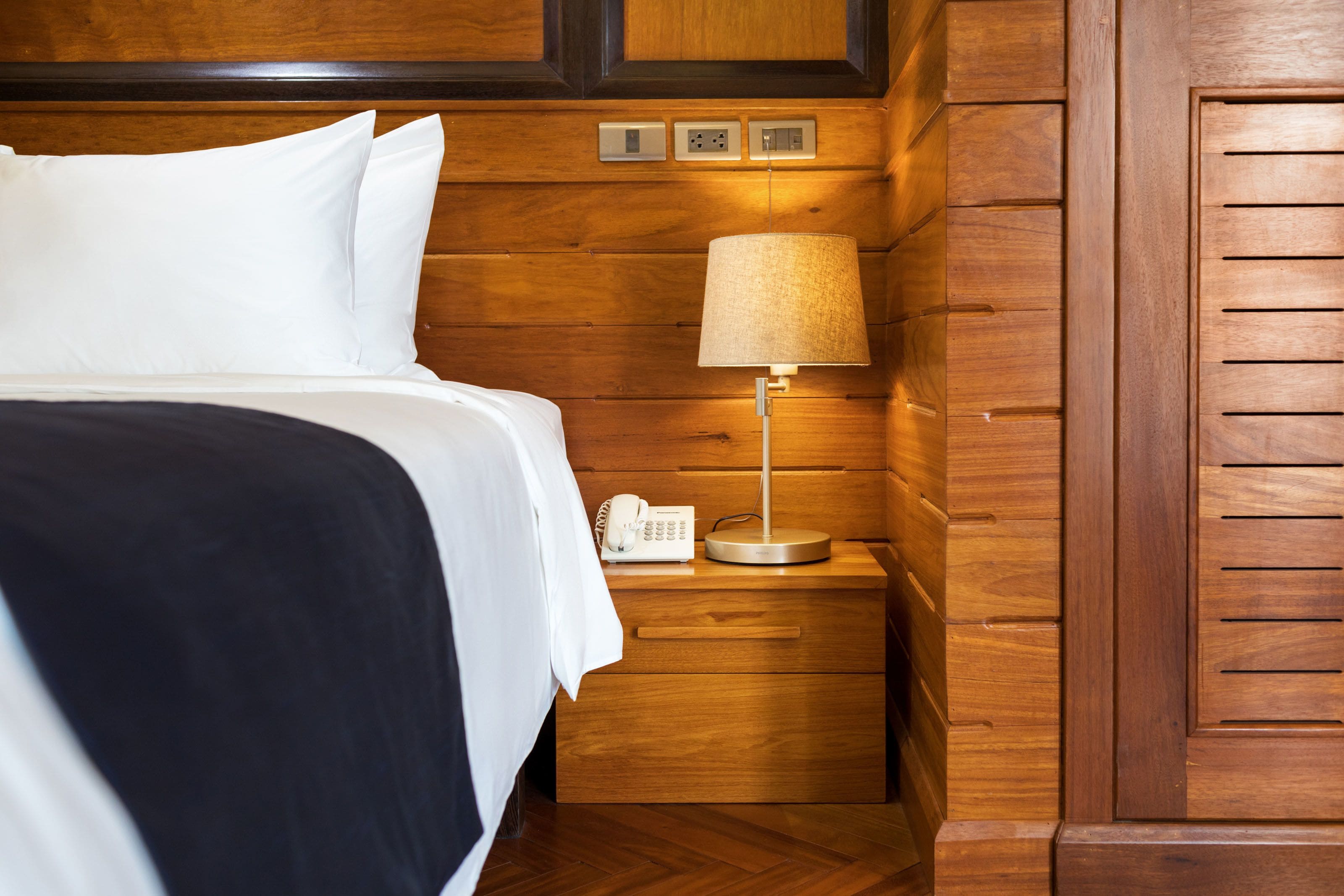 A neatly made bed with white linens, a bedside lamp, and a phone on a wooden nightstand.