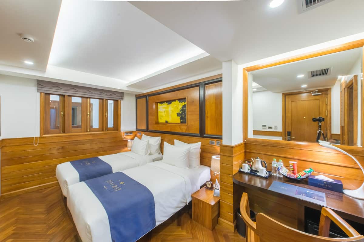 A hotel room with two beds, wooden flooring, a desk with a chair, a mirror, and a wall-mounted artwork.