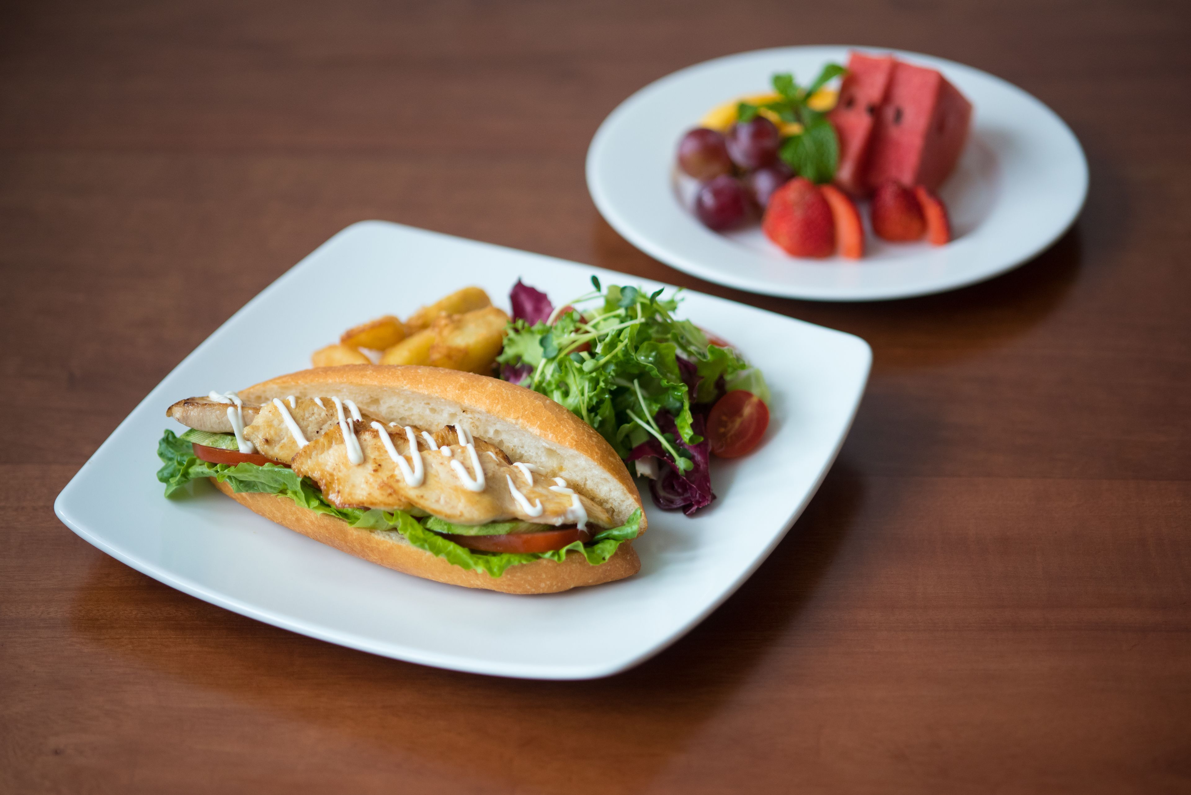 A sandwich on a white plate with a side salad and fruit on another plate.