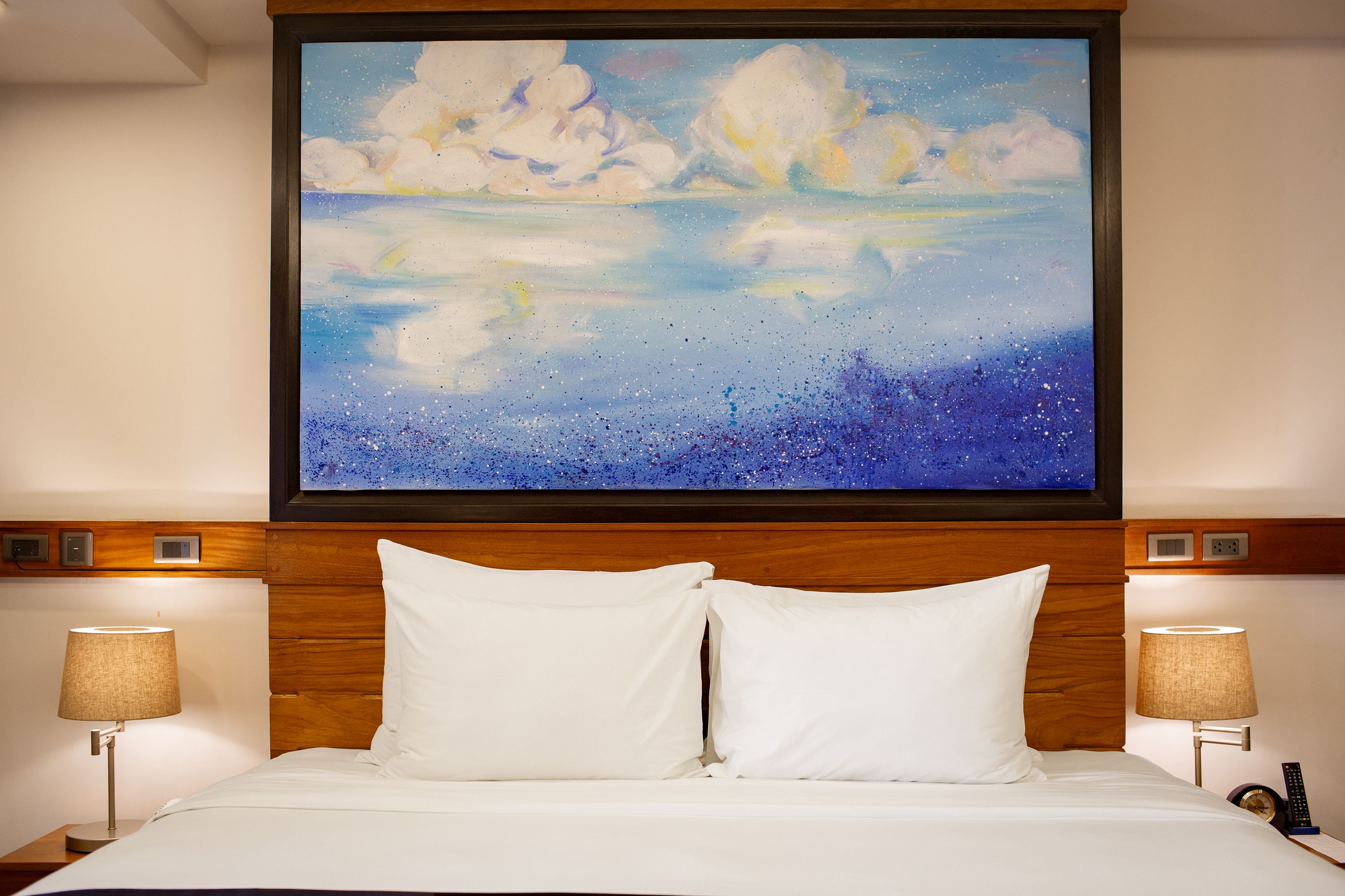 A hotel room with a large painting of clouds and a starry sky above the bed, wooden headboard, two bedside lamps, and a nightstand with a remote control.