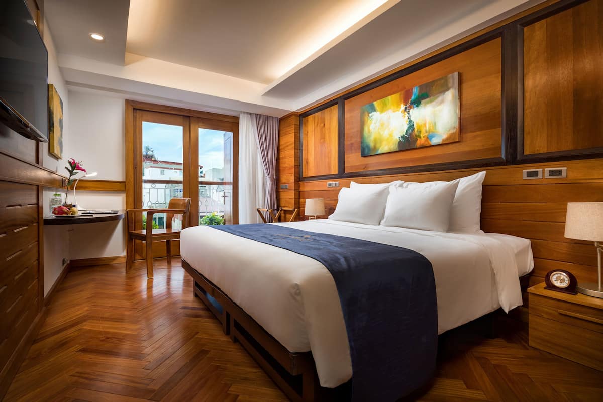 A hotel room with a large bed, wooden paneling, a painting on the wall, a desk with a chair, and a window with a view of buildings.