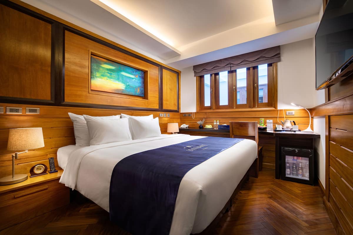 A hotel room with a large bed, wooden paneling, a painting above the bed, a desk with a refrigerator, and a television.