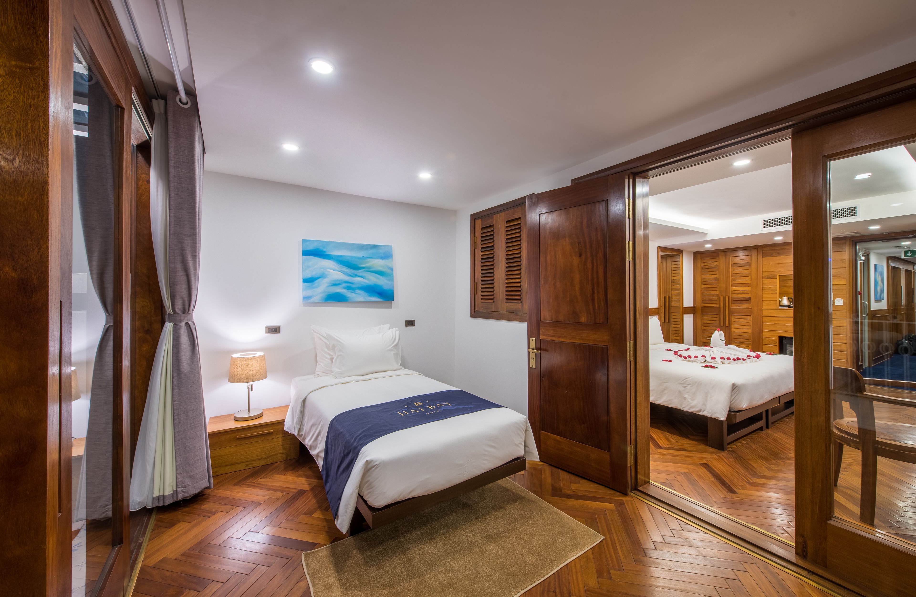 A hotel room with a bed, wooden flooring, a nightstand with a lamp, a painting on the wall, and a view of another room through an open door.