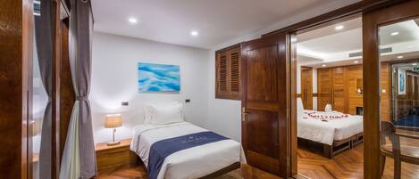 A hotel room with a bed, wooden flooring, a nightstand with a lamp, a painting on the wall, and a view of another room through an open door.