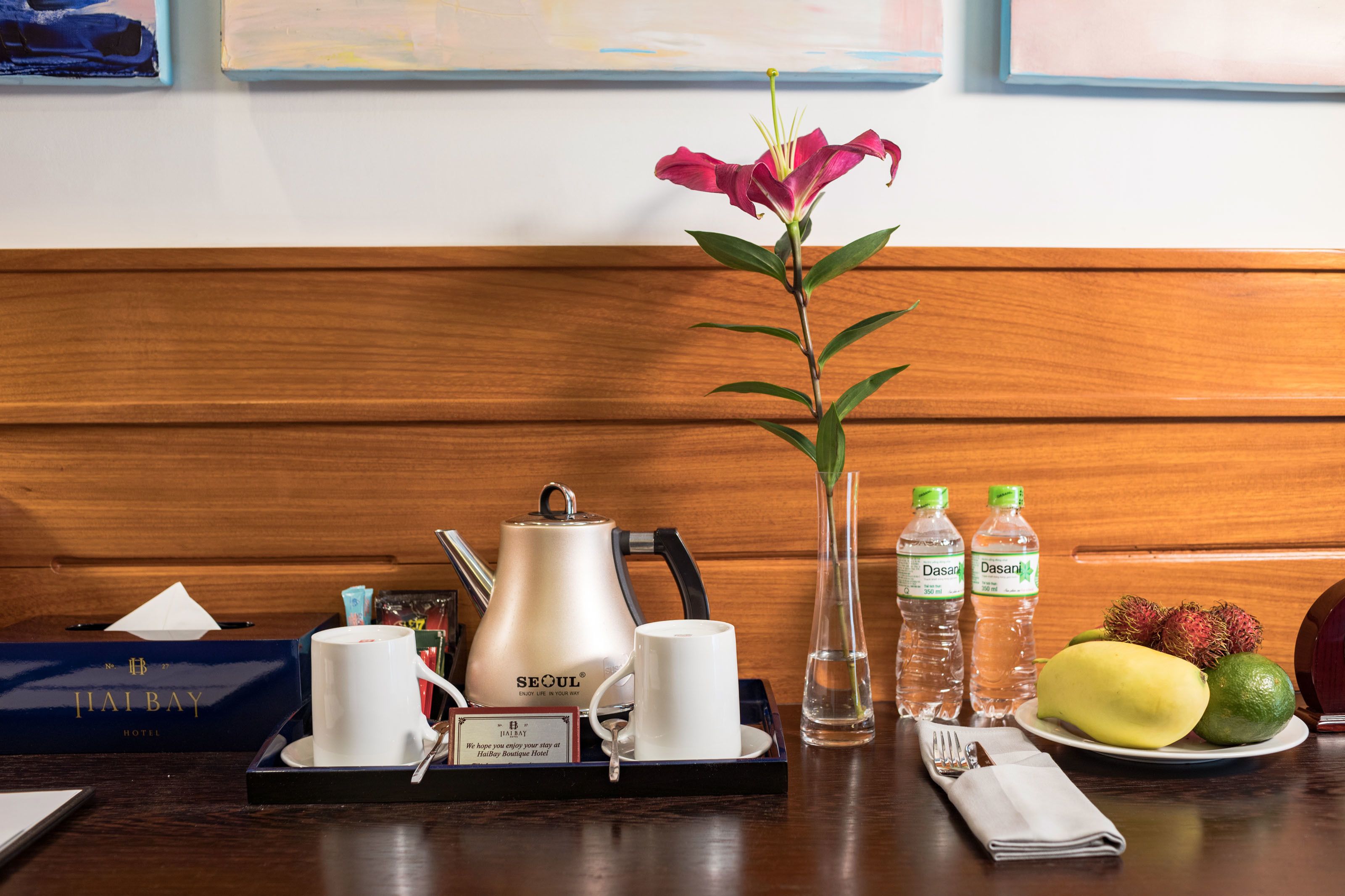 A hotel room with a wooden panel wall, a tray with a teapot, cups, a vase with a flower, bottles of Dasani water, a plate with fruit, and a folded napkin.