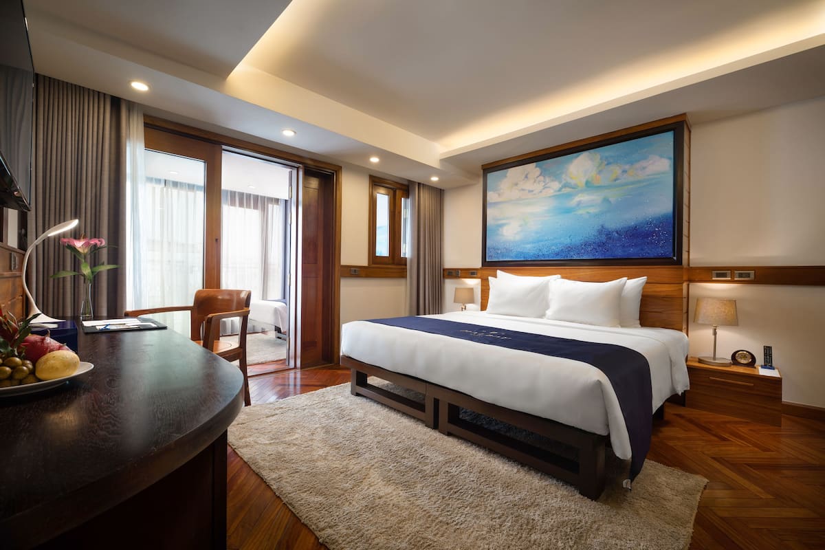 A hotel room with a large bed, a desk with a lamp, a chair, a painting on the wall, and a view of the outdoors.