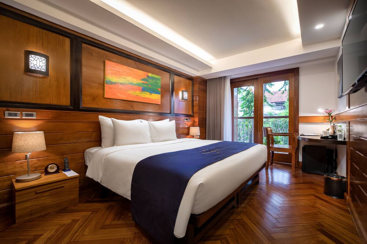 A hotel room with a large bed, wooden paneling, a painting, a desk with a chair, and a TV.