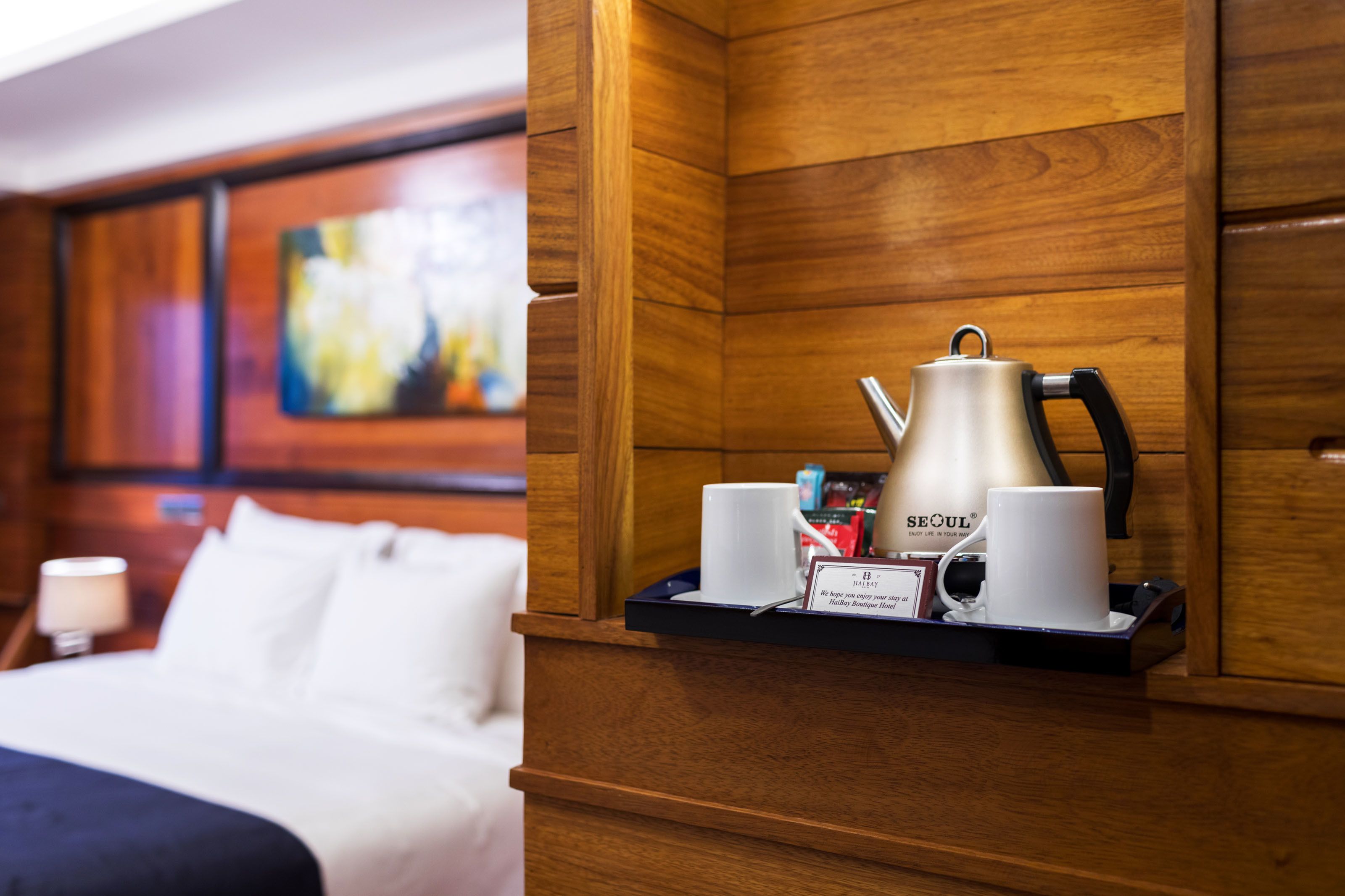 A hotel room with a wooden headboard, a bed with white bedding, a coffee station with a coffee pot, two white cups, and a tray with a sign.