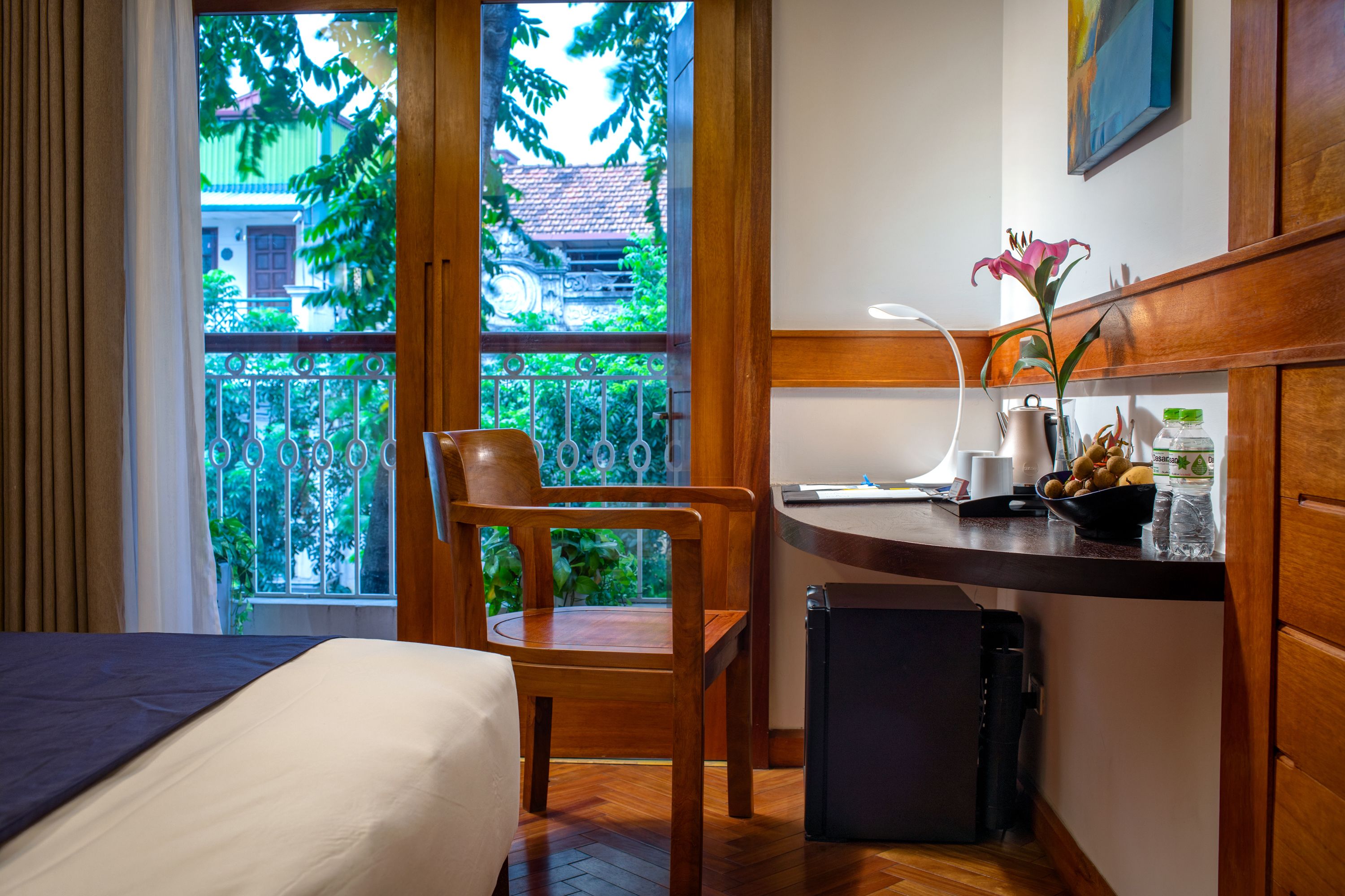 A hotel room with a bed, a wooden chair, a small table with a vase of flowers, a kettle, and a bowl of fruit.
