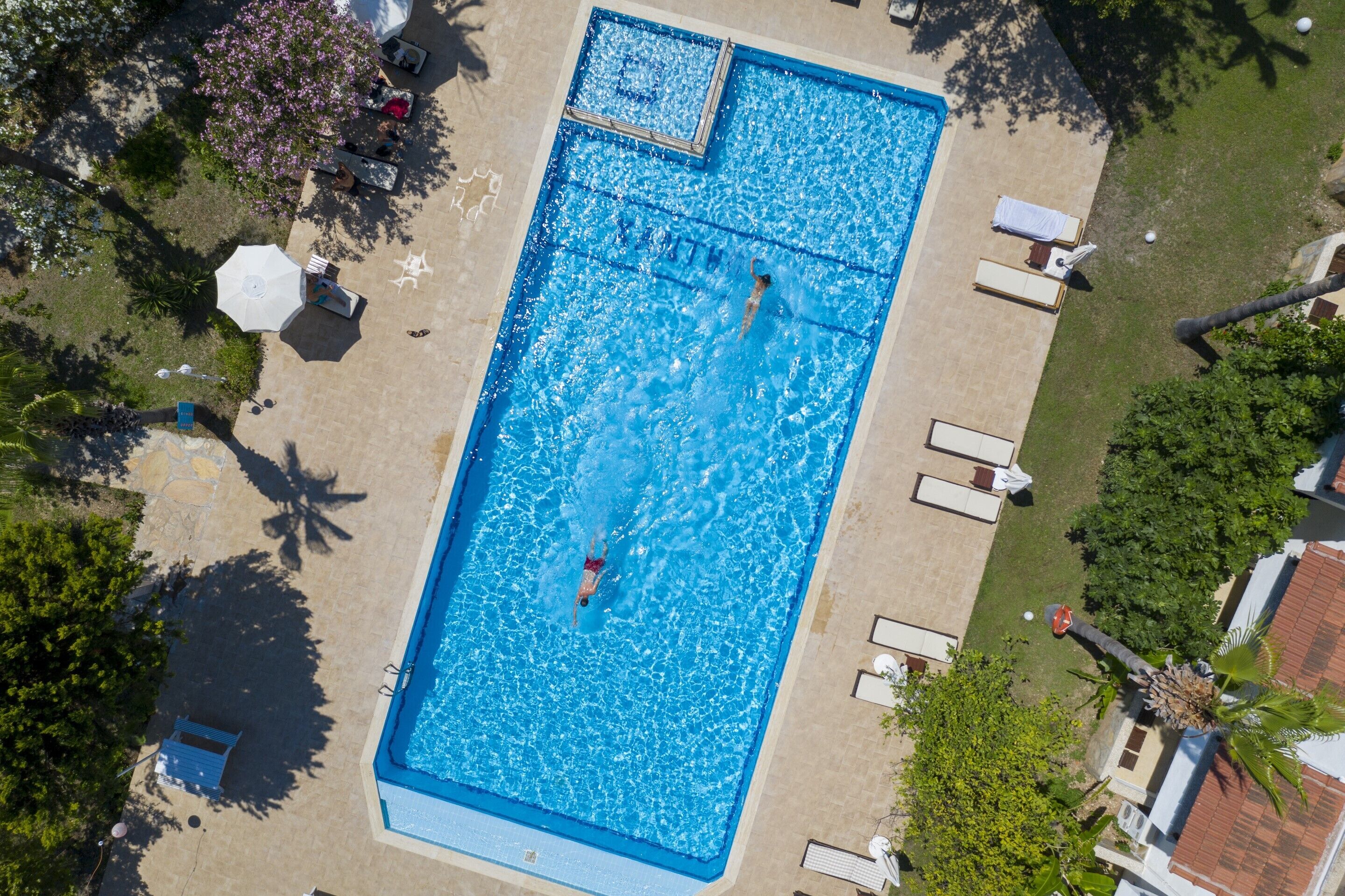 A rectangular swimming pool with a diving board, surrounded by lounge chairs and umbrellas, with trees and a building in the background.