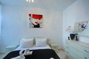 A modern bedroom with a bed, white pillows, a nightstand, a dresser, and a wall with a painting and a mirror.