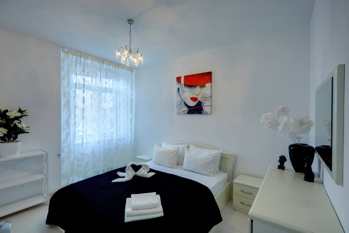 A modern hotel room with a bed, white nightstands, a dresser, a chandelier, a large window with sheer curtains, a painting on the wall, and a potted plant.