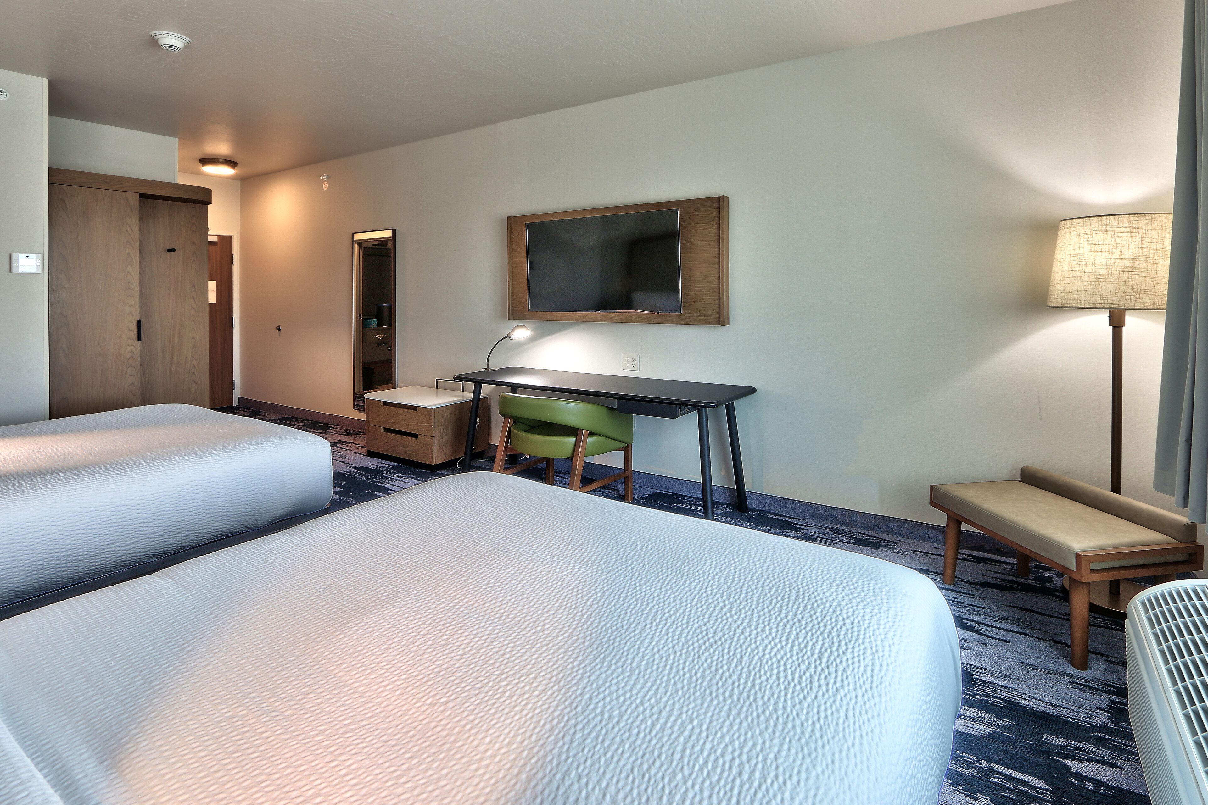 A hotel room with two beds, a desk, a chair, a lamp, and a bench.