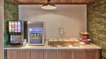 Estación de desayuno autoservicio con dispensador de bebidas, refrigerios y una exhibición de frutas.