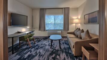 Quarto de hotel moderno com televisão de tela plana, escrivaninha com cadeira, sofá, mesinha de centro e uma janela grande com cortinas.