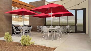 An outdoor patio with metal chairs and tables, red umbrellas, and a barbecue grill.