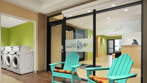 A laundry area with a washing machine, two turquoise Adirondack chairs with colorful cushions, and a reception desk with a sign reading "SPIN Cycle." - Home2 Suites by Hilton Elko (Elko)