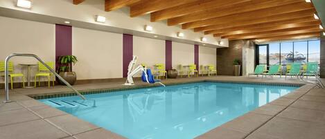 An indoor swimming pool with clear blue water, wooden ceiling beams, and yellow chairs around the pool.