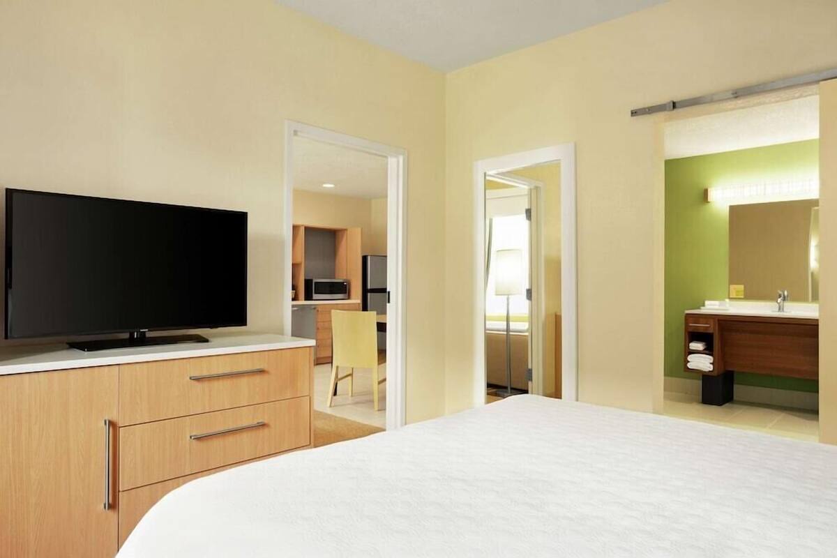 Een hotelkamer met een bed, een televisie op een dressoir, een badkamer met een spiegel en een kitchenette op de achtergrond.