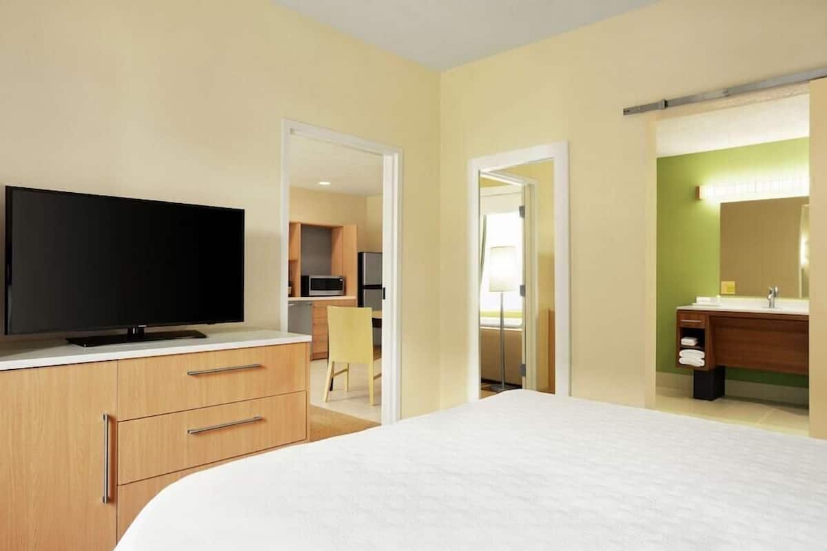 A hotel room with a bed, a TV on a dresser, a bathroom with a mirror, and a kitchenette in the background.