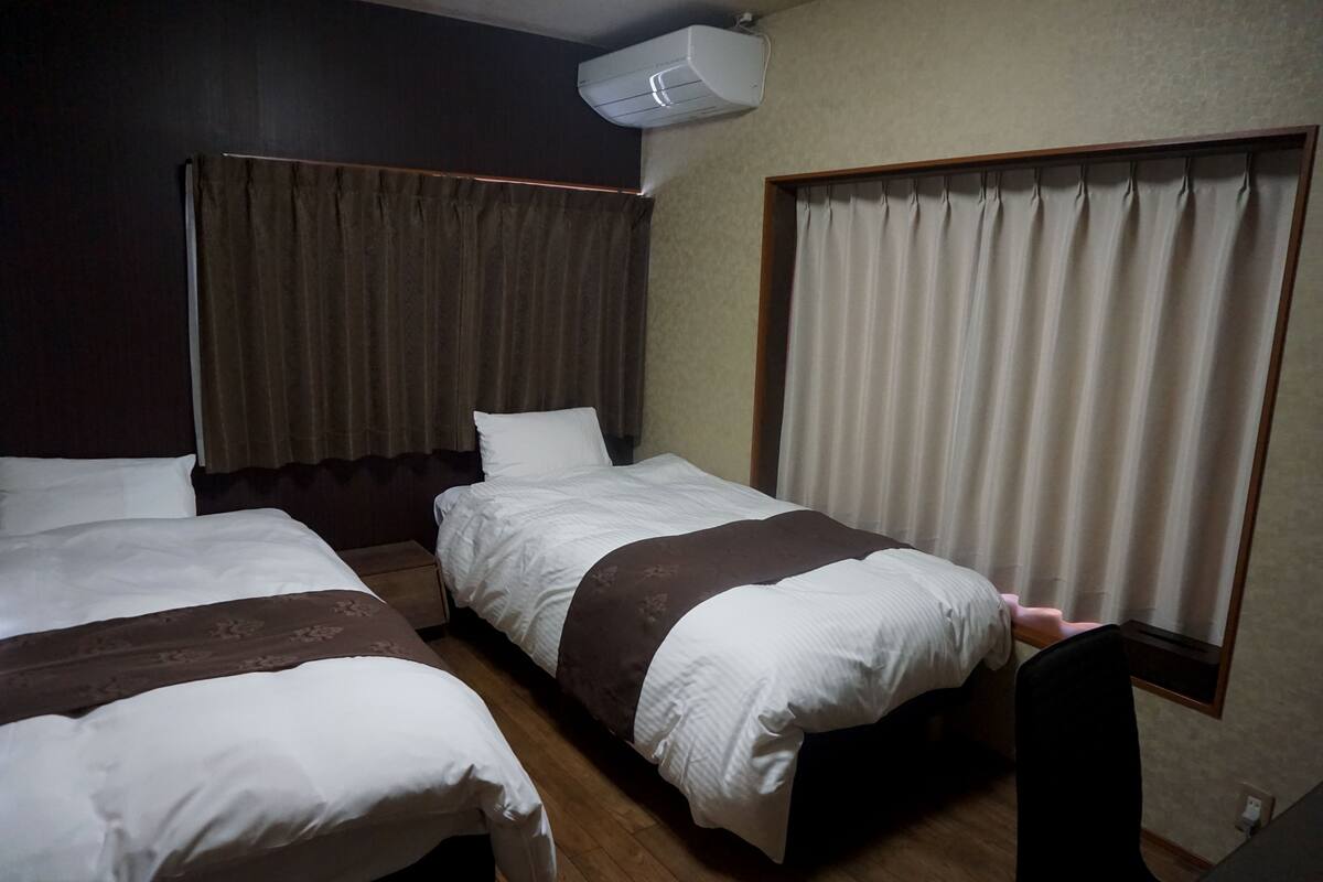 A hotel room with two single beds, a window with curtains, an air conditioning unit, and a wooden floor.