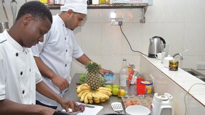 A kitchen with chefs preparing food, featuring a cutting board, bananas, pineapple, and various kitchen utensils. - Chukwani Executive Inn (Zanzibar Town)