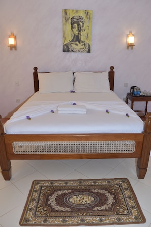 A neatly made bed with white linens and a decorative pillow, a wooden headboard, a wall-mounted painting, and a small table with a lamp and a bottle. - Chukwani Executive Inn (Zanzibar Town)
