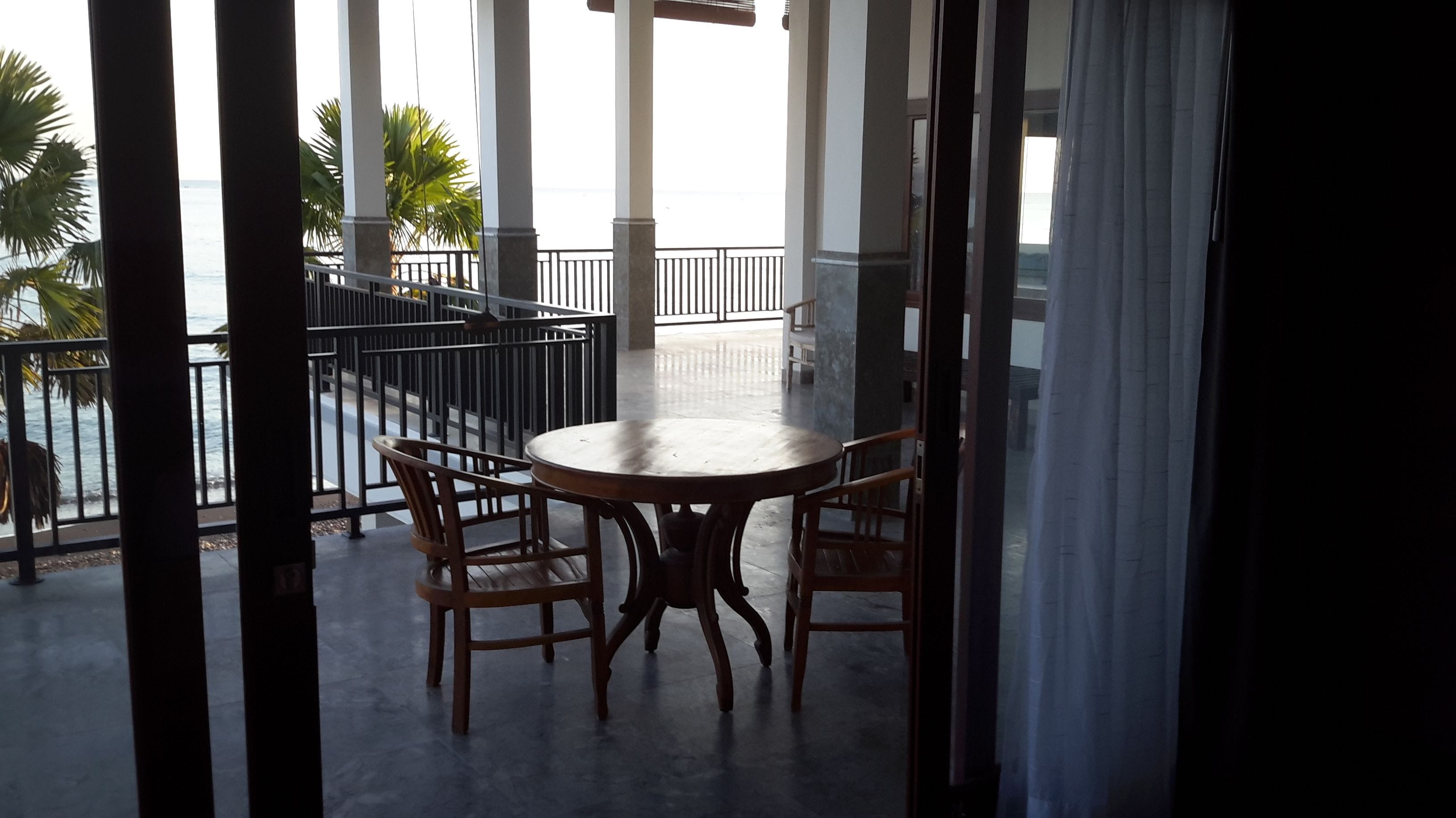A balcony with a round table and two chairs, overlooking a palm tree and a view of the sea.