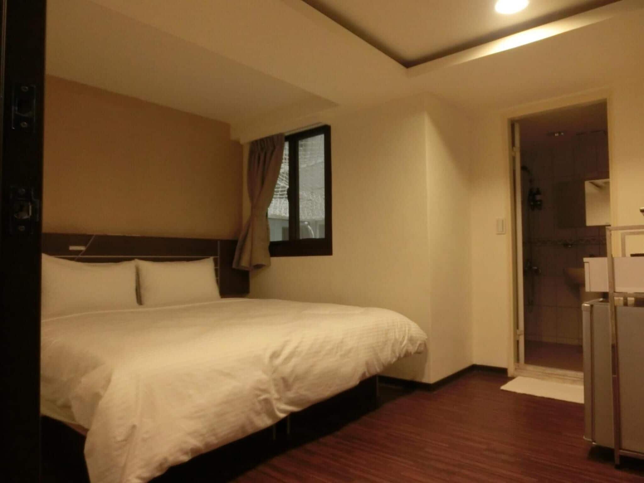A hotel room with a single bed, a small window with curtains, a door leading to a bathroom, and a wooden floor.