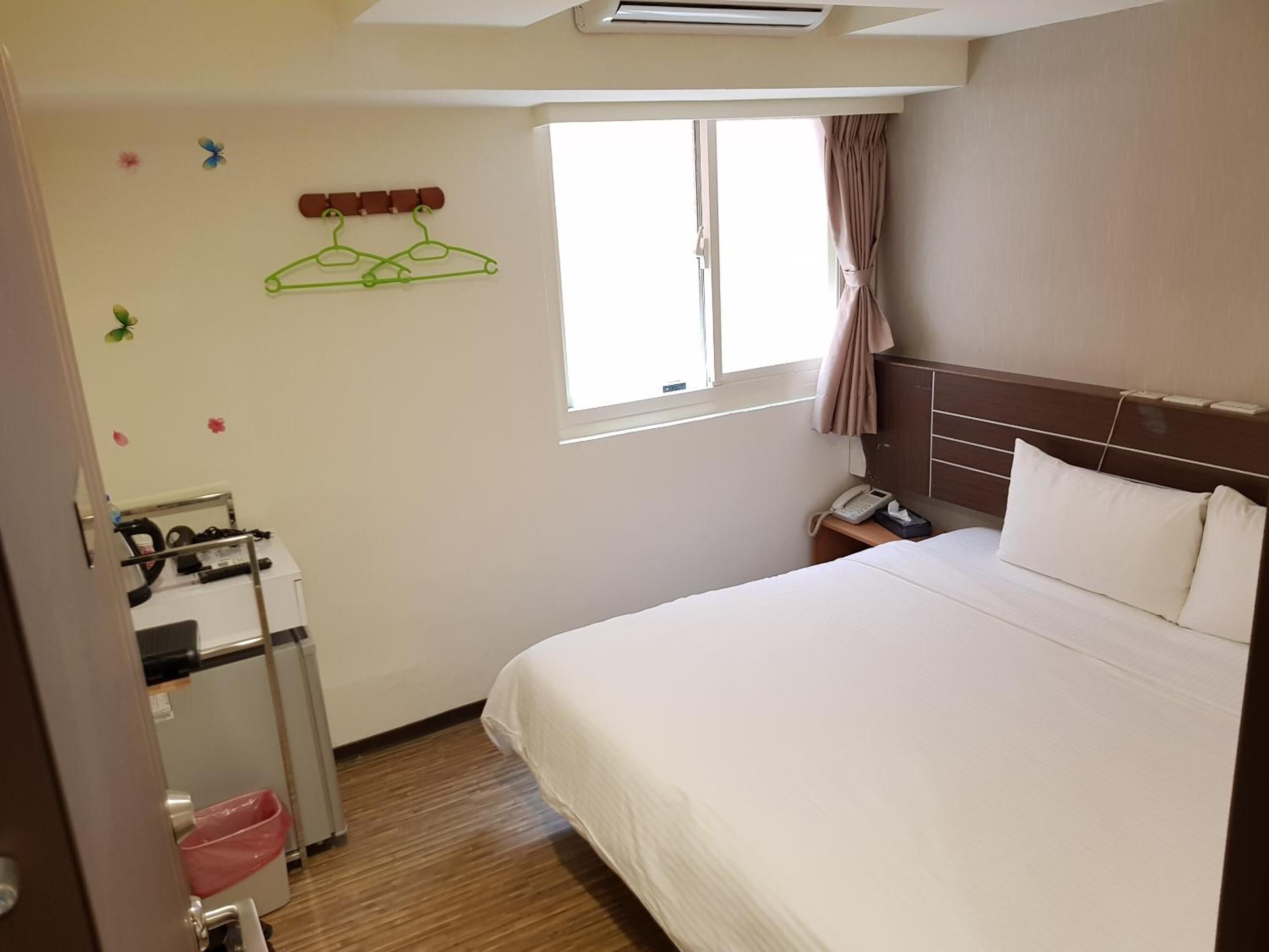 A hotel room with a bed, a nightstand, a telephone, a window with curtains, and a wall-mounted air conditioning unit.